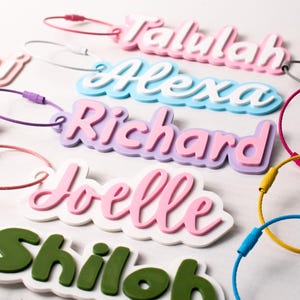 May include: A collection of colorful name tags with various names in a playful font. The tags are in shades of pink, blue, green, and purple, with matching or contrasting colored loops for attachment. The names include "Talulah", "Alexa", "Richard", "Joelle", and "Shiloh".