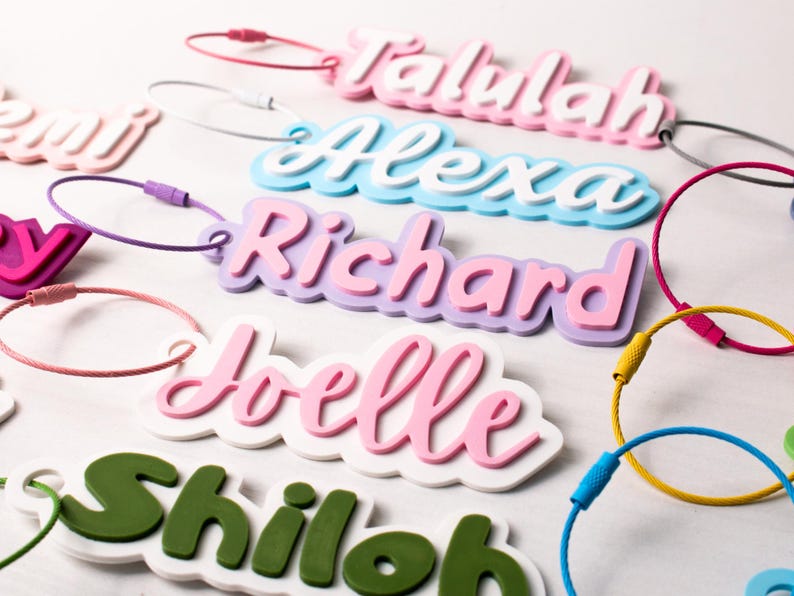 May include: A collection of colorful name tags with various names in a playful font. The tags are in shades of pink, blue, green, and purple, with matching or contrasting colored loops for attachment. The names include "Talulah", "Alexa", "Richard", "Joelle", and "Shiloh".