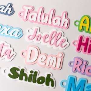 May include: A collection of colorful, layered name cutouts in various fonts. Names include Talulah, Alexa, Joelle, and others, in shades of pink, blue, green, and purple. Each cutout has a small hole for hanging or attaching.