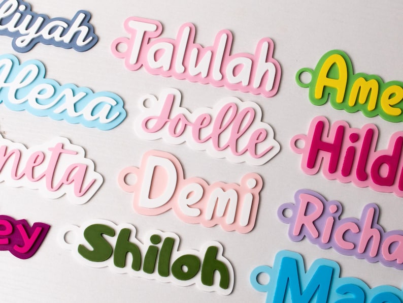 May include: A collection of colorful, layered name cutouts in various fonts. Names include Talulah, Alexa, Joelle, and others, in shades of pink, blue, green, and purple. Each cutout has a small hole for hanging or attaching.