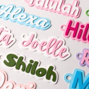May include: Close-up of colorful name cutouts in various fonts and colors. Names include Alexa, Joelle, and Shiloh. Each name is layered with a white outline and a small hole for hanging. The names are in pink, blue, green, and purple.