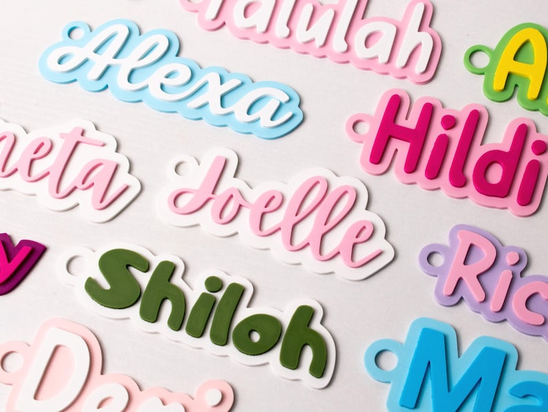 May include: Close-up of colorful name cutouts in various fonts and colors. Names include Alexa, Joelle, and Shiloh. Each name is layered with a white outline and a small hole for hanging. The names are in pink, blue, green, and purple.