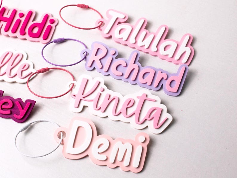 May include: A collection of personalized name tags in various colors. Each tag features a different name in a layered design with a matching loop for attachment. The names include "Hildi", "Talulah", "Richard", "Kineta", and "Demi".