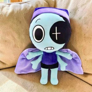 Puede incluir: Una mu&ntilde;eca de peluche con una cara azul claro, un ojo negro grande y un parche en el ojo negro con una cruz blanca. La mu&ntilde;eca tiene una capa, un sombrero y una camisa morados, con pantalones cortos negros y brazos y piernas azul claro. La mu&ntilde;eca est&aacute; en un sof&aacute; beige.