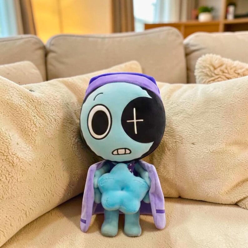 Puede incluir: Una mu&ntilde;eca de peluche con una cara azul claro, un parche en el ojo de media luna negro y una gorra y capa moradas. La mu&ntilde;eca sostiene una estrella azul y tiene una boca cosida. La mu&ntilde;eca est&aacute; sentada en un sof&aacute; beige.