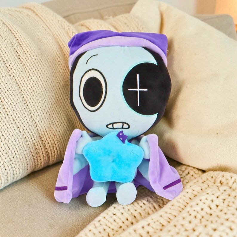 Puede incluir: Una mu&ntilde;eca de peluche con una cara azul claro, un ojo negro grande y un parche negro con una cruz blanca. Lleva un gorro de dormir y una capa morados, y sostiene una estrella azul. La mu&ntilde;eca est&aacute; dise&ntilde;ada para parecerse a un personaje de dibujos animados.