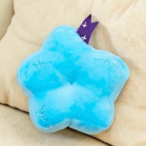 Puede incluir: Un juguete de peluche en forma de estrella azul claro con un aspecto suave y similar a una nube. El juguete tiene detalles cosidos y una cinta morada con cruces blancas. El juguete est&aacute; sobre una superficie beige.