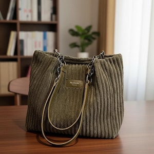 May include: Olive green corduroy tote bag with silver chain handles and olive green leather straps. The bag has a rectangular shape and a small rectangular tag that reads "Mekluola 1932". The bag is sitting on a wooden surface.