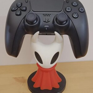 Puede incluir: Un mando de videojuego negro apoyado sobre un soporte con forma de personaje blanco y rojo. El soporte presenta una cabeza blanca con dos cuernos y ojos ovalados negros, con una capa roja, montado sobre una base circular de color negro.