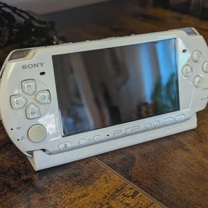 May include: A white Sony PSP handheld gaming console, featuring a screen and control buttons. The console is resting on a white stand, with the Sony logo visible. The background is a wooden surface.