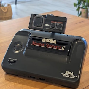 Puede incluir: Una consola de videojuegos Sega Master System II negra con un mando. La consola tiene el texto "SEGA Master System II" en rojo y blanco. El mando es negro con una cruceta y botones. La consola está sobre una superficie de madera.