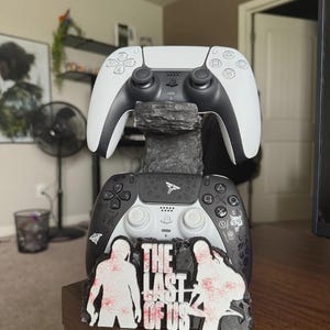 May include: A dual controller stand featuring a rock-like texture, holding a white and black game controller on top and a black controller with white symbols and The Last of Us logo on the base, which depicts two silhouettes and red splatter.