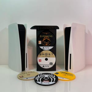 Könnte beinhalten: Eine Präsentation mit einem schwarzen Spielhalter mit zwei weißen und schwarzen Spielkonsolen. Der Halter enthält zwei Spiel-Discs mit den Aufschriften "Elden Ring" und "Ghost of Tsushima". Drei weitere Spiel-Discs sind sichtbar.