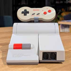 Puede incluir: Una consola gris del sistema de entretenimiento de Nintendo con un mando a juego. La consola tiene un botón de encendido rojo y el texto "Nintendo Entertainment System". El mando tiene una cruceta, select, start y botones rojos.