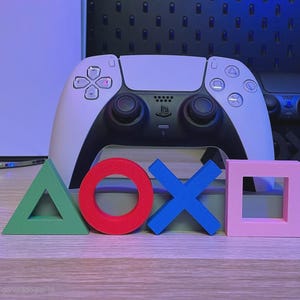 May include: A white and black wireless game controller positioned behind four distinct 3D symbols: a green triangle, a red circle, a blue cross, and a pink square. The controller features a central black touchpad, dual analog sticks, and standard action buttons.