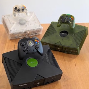 May include: Three Xbox consoles in the image. One is black, one is clear, and the other is olive green. Each console has a matching controller on top. The black console has a green Xbox logo. The controllers have colored buttons.