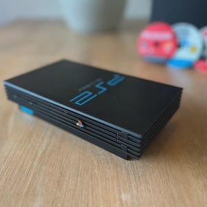 Puede incluir: Una consola PlayStation 2 negra con el texto "PS2" en azul. La consola es rectangular con una ranura para discos y un pequeño logotipo colorido. La consola está sobre una superficie de madera.
