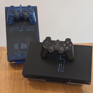 May include: Two black PlayStation 2 consoles with matching controllers. One console is upright and blue, with a blue controller on top. The other console is black with a black controller on top. The PS2 logo is visible on both consoles.