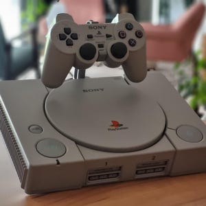 Peut inclure: Une console PlayStation grise avec une manette assortie. La console dispose d'un lecteur de disque, de boutons d'alimentation et de réinitialisation, et de deux emplacements pour cartes mémoire. La manette a un pavé directionnel, des boutons d'action et des boutons d'épaule.