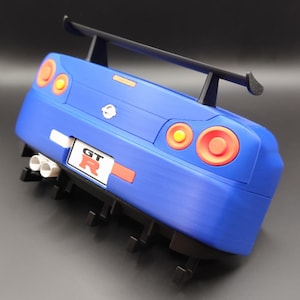 May include: A blue toy car with a black spoiler and a GT R license plate. The car has orange and red taillights and headlights. The car has white exhaust pipes and black fins on the bottom.