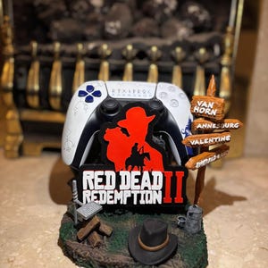 May include: A white video game controller stand featuring a Red Dead Redemption II logo, a black cowboy hat, a small campfire, and a wooden signpost with the names Van Horn, Annesburg, Valentine, and Emerald Ranch printed on the individual boards.