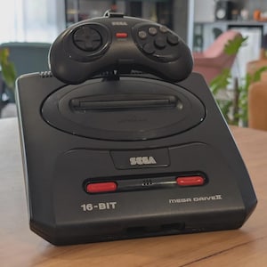 May include: A black Sega Genesis console with a matching controller on top. The console has red accents and the text "16-BIT" and "MEGA DRIVE II". The controller has a directional pad and several buttons.