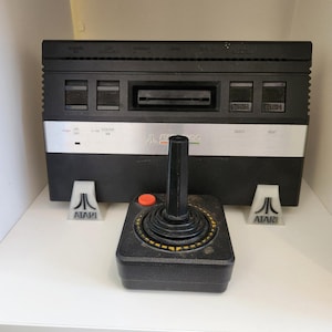 以下が含まれることがあります： 黒とシルバーのヴィンテージAtariビデオゲームコンソール。オレンジ色のボタンが付いたジョイスティックと、Atariロゴのスタンドが2つあります。コンソールには、カートリッジスロットといくつかのボタンがあります。