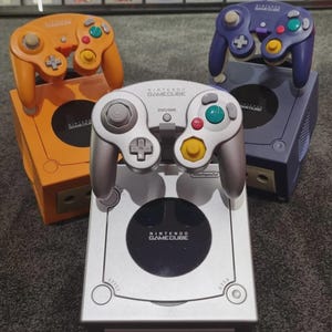 Könnte beinhalten: Silberne, orangefarbene und violette Nintendo GameCube Konsolen und Controller. Die Controller haben farbige Tasten und Richtungspads. Die Konsolen sind auf einer grauen Oberfläche gestapelt. Das GameCube-Logo ist sichtbar.