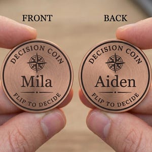 May include: Two circular copper-toned decision coins featuring a brushed metal finish. Each coin displays a compass icon, the text 'DECISION COIN' and 'FLIP TO DECIDE' in black, with the names 'Mila' on the left coin and 'Aiden' on the right coin.