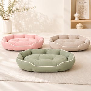 May include: Three pet beds in soft pastel colors: one light pink, one beige, and one sage green. Each bed features a tufted, cushioned center, raised padded sides for support, and a thin white piped trim along the top edge of the bolster.