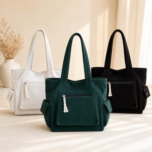 May include: Three corduroy tote bags in white, dark green, and black. Each bag has a front zippered pocket, two side pockets, and a matching tassel zipper pull. The bags have sturdy handles for carrying.