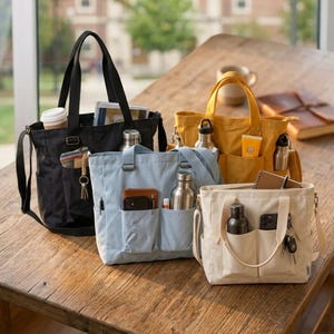 May include: Four canvas tote bags in black, light blue, yellow, and off-white, each with multiple pockets and carrying handles. The bags are filled with items like water bottles, notebooks, and keys, and are displayed on a wooden table.