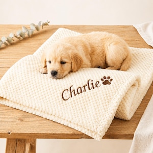 Puede incluir: Un cachorro de color beige claro descansa sobre una manta crema texturizada. La manta está bordada con el nombre "Charlie" en escritura marrón, junto con una huella de pata. El cachorro tiene pelaje marrón claro.