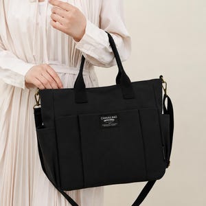 May include: Black canvas bag with multiple pockets and a shoulder strap. The bag has two top handles and a label that reads "CANVAS BAG". The bag is held by a person wearing a cream-colored dress.