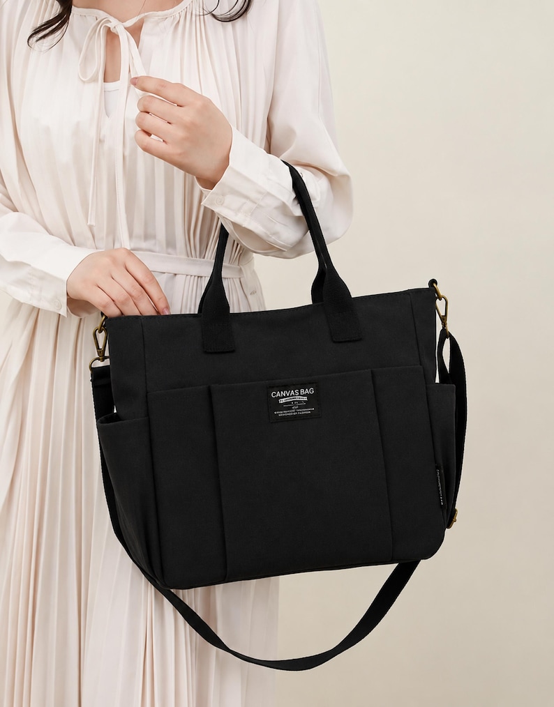 May include: Black canvas bag with multiple pockets and a shoulder strap. The bag has two top handles and a label that reads "CANVAS BAG". The bag is held by a person wearing a cream-colored dress.