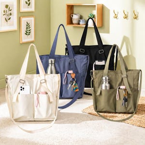 Può includere: Quattro borse tote in tela color crema, blu navy, nero e verde oliva. Ogni borsa ha una tasca frontale, una tasca laterale e un portabottiglie. Le borse hanno tracolle e manici superiori. Chiavi e telefoni sono visibili.