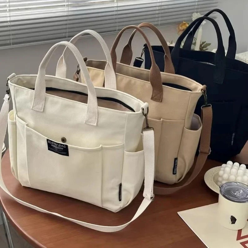 May include: Three canvas tote bags in cream, tan, and black. Each bag has a top handle, a detachable shoulder strap, and multiple exterior pockets. The cream bag has a label that reads "CLOTH BAG". The bags are on a wooden table.