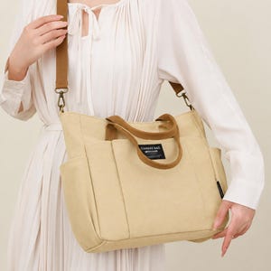 May include: Beige canvas tote bag with brown accents. Features a shoulder strap and top handles. The bag has multiple pockets and a label that reads "CANVAS BAG".