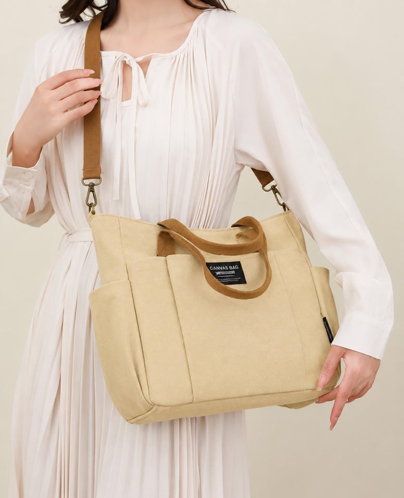 May include: Beige canvas tote bag with brown accents. Features a shoulder strap and top handles. The bag has multiple pockets and a label that reads "CANVAS BAG".