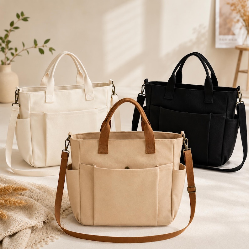 May include: Three canvas tote bags in cream, tan, and black, each with a top handle and a detachable shoulder strap. The bags have multiple exterior pockets and brown leather-like accents on the handles and straps.