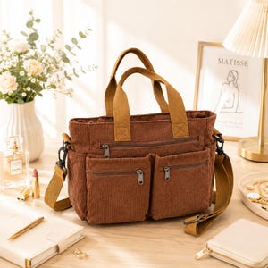 Op de afbeelding: Een bruine corduroy tote bag met meerdere ritsvakken en een schouderriem. De tas heeft twee handvatten aan de bovenkant en een voorvak. De tas staat op een houten oppervlak.