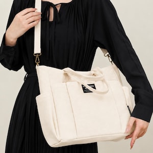 May include: Cream-colored tote bag with a shoulder strap and multiple pockets. The bag has a rectangular shape with a top handle and a small label on the front. The strap is a lighter cream color and is attached with gold-colored hardware.