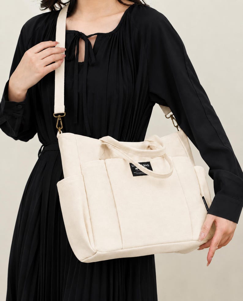 May include: Cream-colored tote bag with a shoulder strap and multiple pockets. The bag has a rectangular shape with a top handle and a small label on the front. The strap is a lighter cream color and is attached with gold-colored hardware.