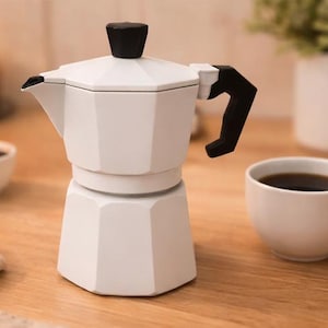 Puede incluir: Una taza de cerámica blanca con asa negra y el texto "But First, Coffee" en negrita negra. La taza se encuentra sobre un fondo borroso, sugiriendo un ambiente acogedor.