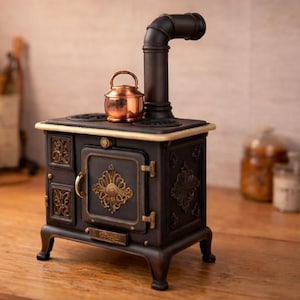 Puede incluir: Una estufa en miniatura negra y dorada con una tetera de cobre encima. La estufa tiene detalles ornamentados y una chimenea. La estufa está sobre una superficie de madera.