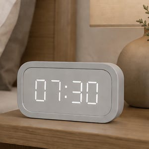 May include: A rectangular, light gray digital alarm clock displaying the time 07:30 in white LED numerals. The clock has rounded corners and sits on a wooden surface, suggesting a bedside table setting.