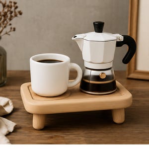 Pode incluir: Uma caneca de cerâmica branca cheia de café escuro está sobre uma pequena bandeja de madeira clara, ao lado de uma cafeteira italiana branca e prateada. A cafeteira italiana tem uma alça e uma tampa pretas. A bandeja tem pernas curtas e arredondadas.