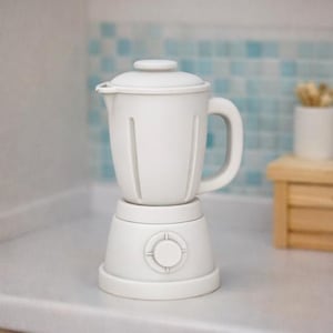 May include: A white blender with a handle, lid, and a base with a circular design. The blender has vertical lines on the pitcher. The background features a light blue and white tile pattern and a small wooden table.
