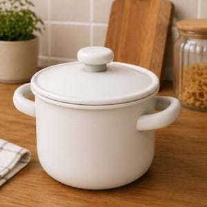 Pode incluir: Uma panela de cozinha esmaltada branca com tampa e duas alças laterais. A panela está sobre uma superfície de madeira. Uma tábua de corte de madeira e um frasco de vidro estão no fundo.