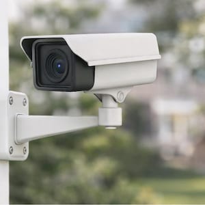 Puede incluir: Cámara de seguridad blanca montada en una pared. La cámara tiene una lente negra y está encerrada en una carcasa blanca con una visera protectora. Montada sobre un brazo y soporte blancos.